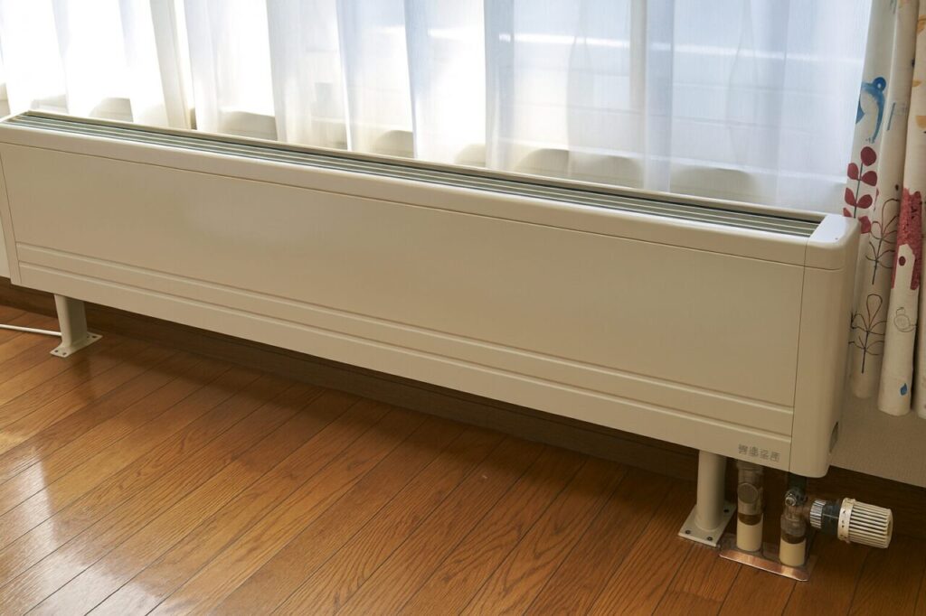 室内の木製フローリングの上に設置されたベージュのパネルヒーター。窓から自然光が差し込み、カーテンの横に静かに配置されている様子。