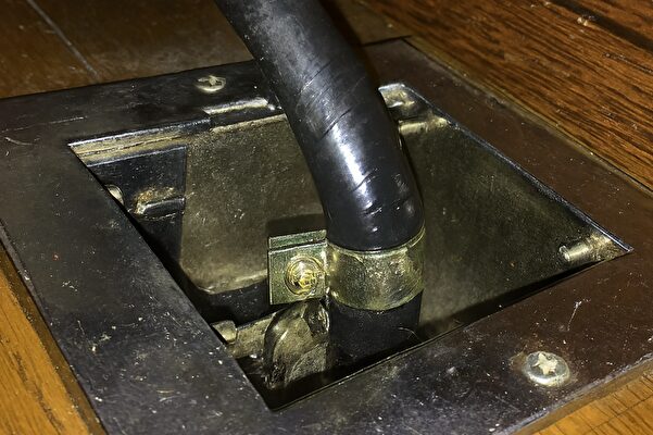 木製の床に埋め込まれた金属プレートの穴から黒いホースが立ち上がっている様子。固定金具でしっかりと締め付けられている。