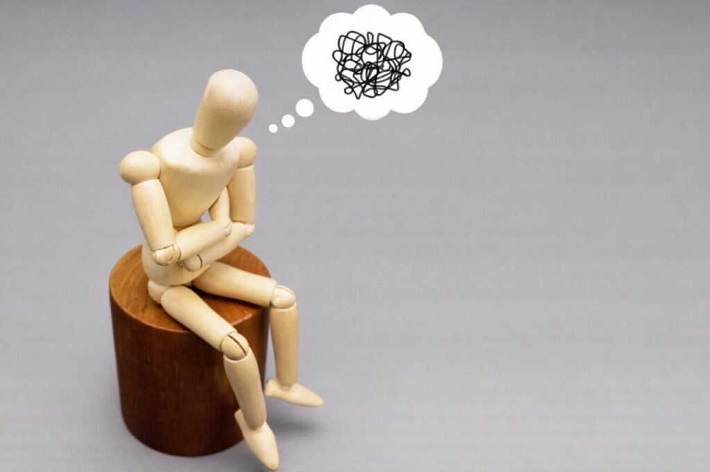 木製のデッサン人形が丸い台に座り、腕を組んで悩みながら頭上にぐちゃぐちゃした思考の吹き出しを浮かべている様子
