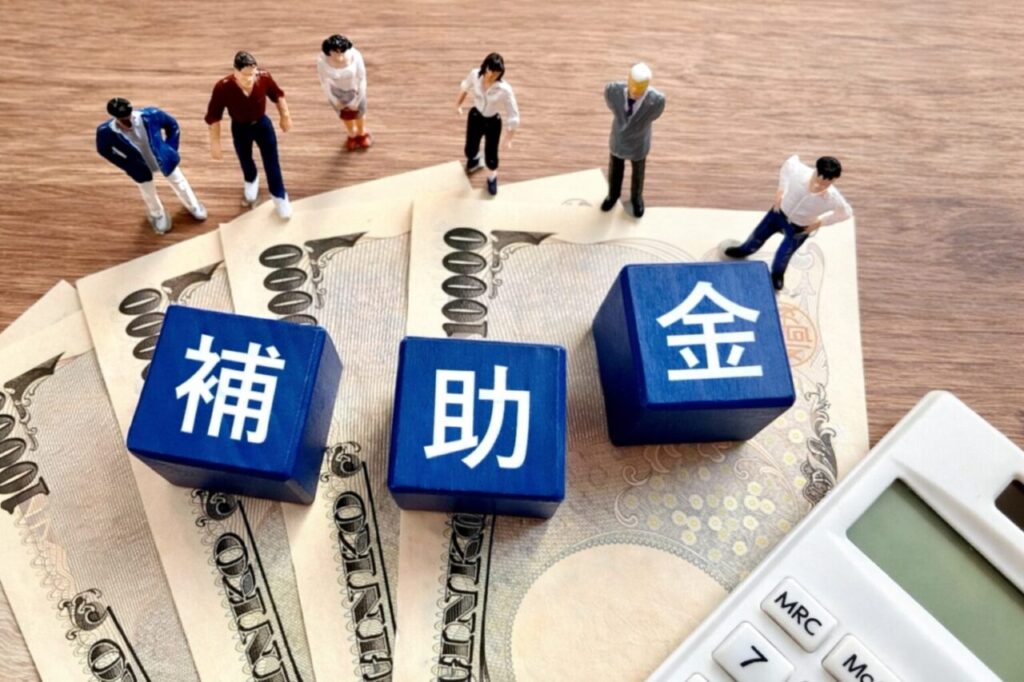補助金を表す青いキューブと一万円札の上に立つ複数の人物模型と電卓のイメージ