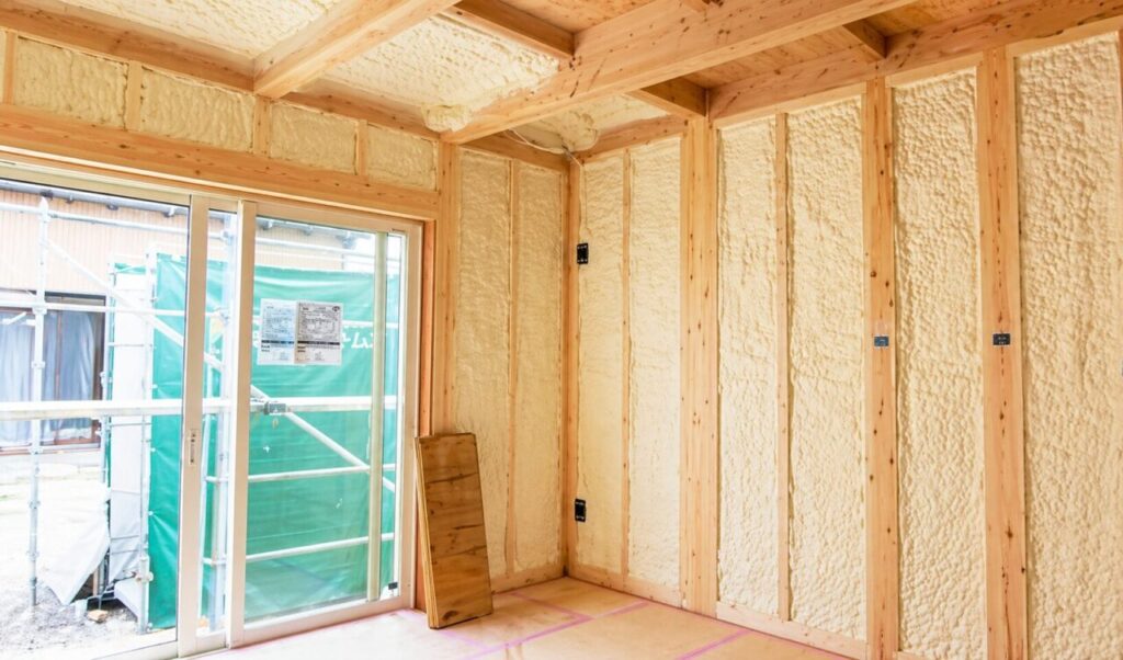 木造住宅の室内で、柱と梁が見える状態に発泡ウレタン断熱材が施工され、掃き出し窓の外に足場と養生シートが見える工事中の空間