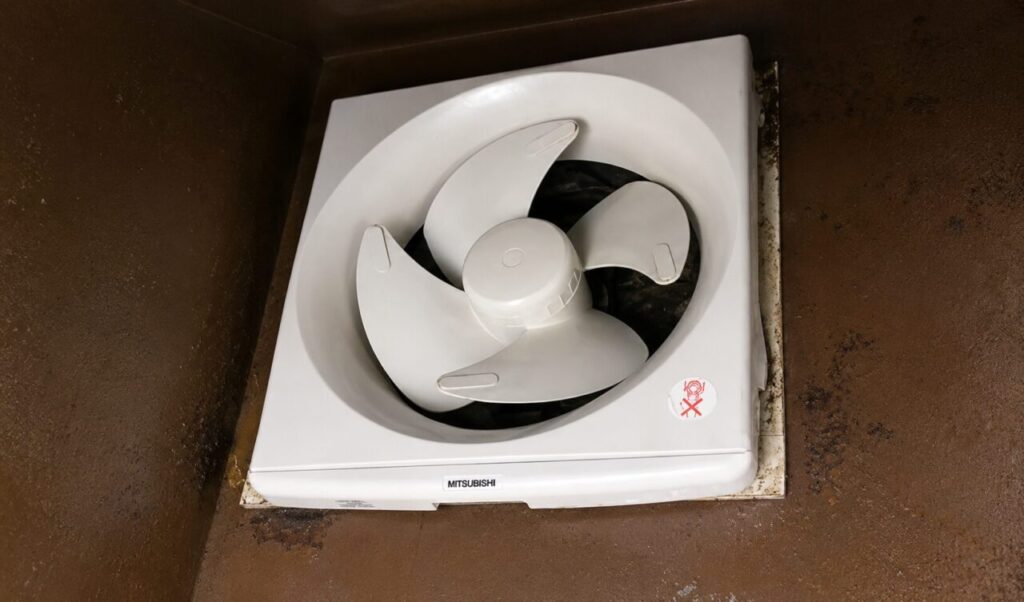 壁面に設置された白色のプロペラ式換気扇を下から見上げた様子。周囲の茶色い換気スペースには汚れや経年劣化が見られ、換気扇本体と取り付け枠の状態が確認できる。