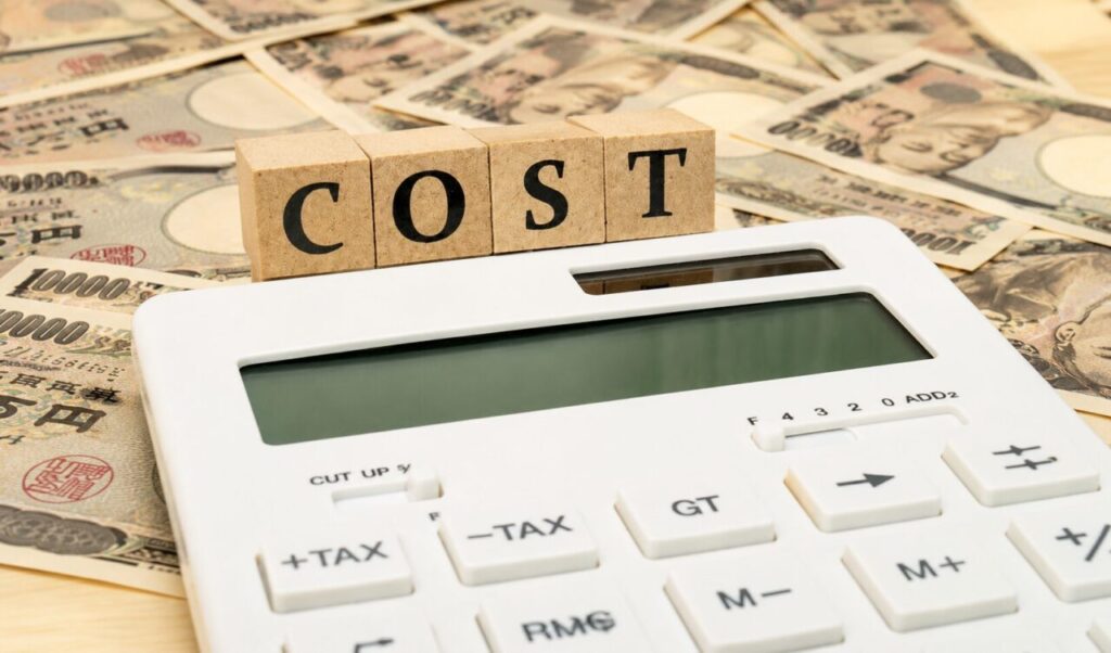 「電卓と『COST』の木製ブロック、背景に広がる日本円紙幣でコストや費用負担をイメージした横長画像」