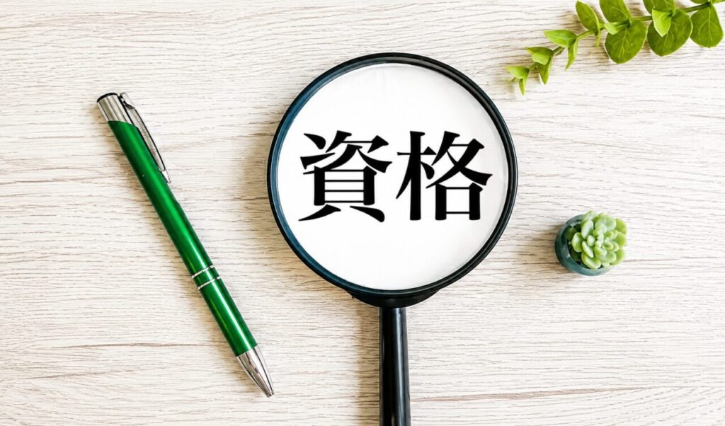 拡大鏡に「資格」の文字が映り、木目調のデスクに緑色のペンと観葉植物が配置された資格取得・試験のイメージ画像。