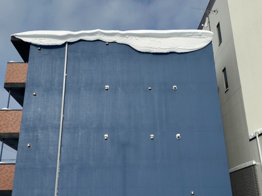 建物の外壁を正面から撮影した様子。屋根の縁に雪が大きく張り出し、雪庇が形成されている状態で、外壁には複数の給排気口が設置されているのが確認できる。