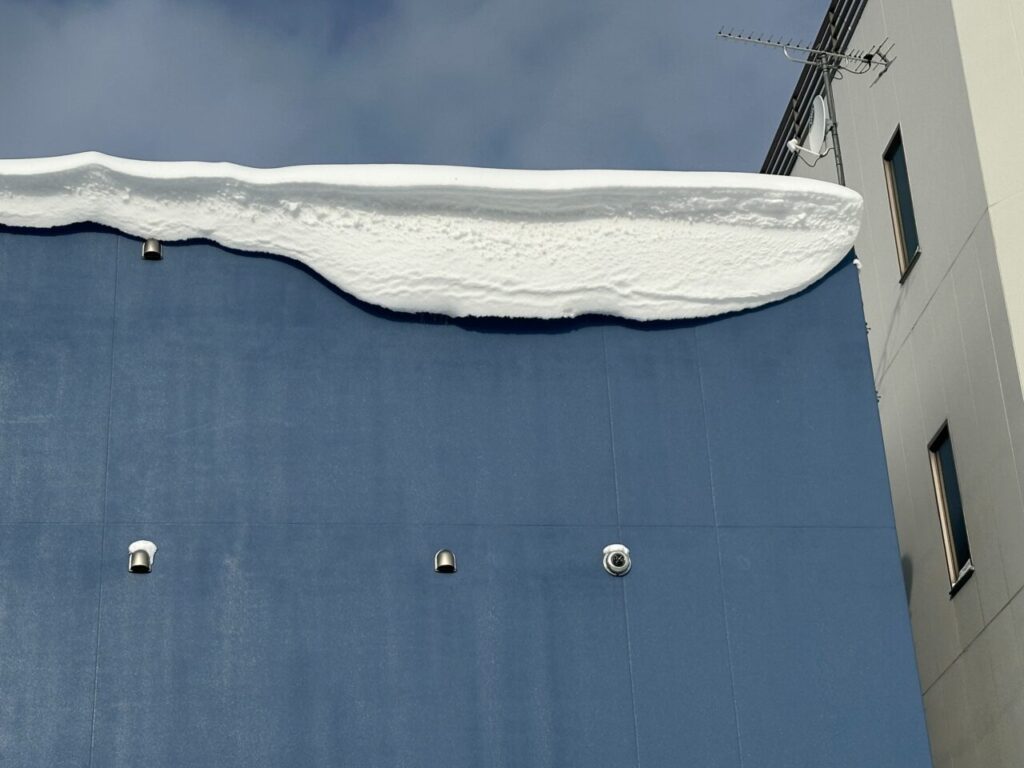 建物外壁の上部を撮影した様子。屋根の縁に厚く積もった雪が張り出し、雪庇が形成されており、外壁には給排気口や配管が設置されている状態。