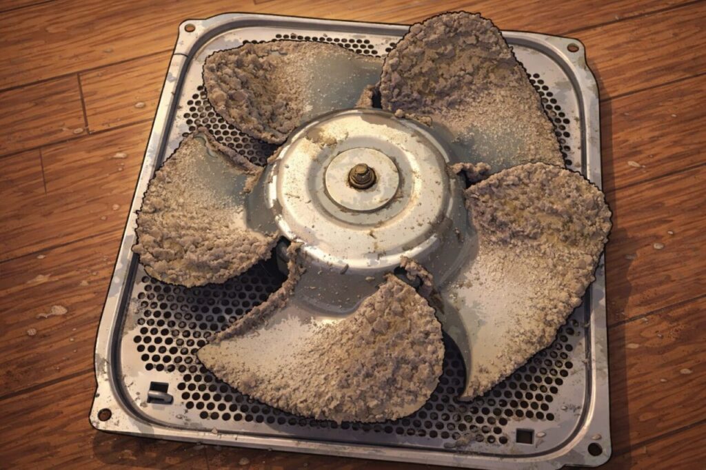 埃が厚く付着して固着した換気扇の羽根と、金属製カバー全体に汚れが広がっている状態をイラスト風で表現した様子