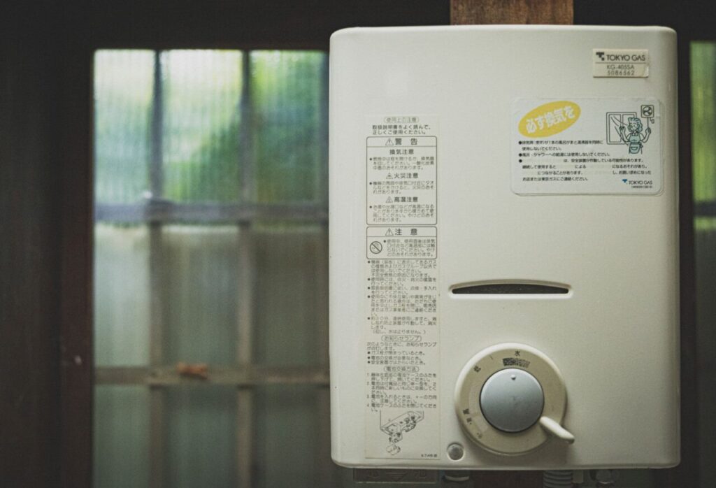 室内の壁に設置された屋内用ガス瞬間湯沸かし器を正面から撮影した様子で、前面には注意書きラベルと温度調整用のダイヤルがあり、背後にはすりガラスの窓が見える設置環境の状態