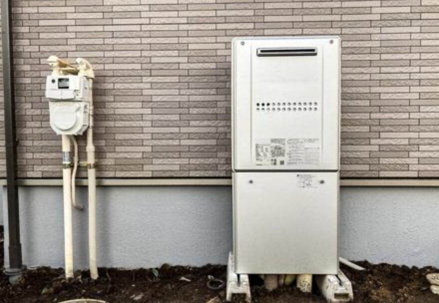 住宅外壁の前に設置された据え置き型のガス給湯器と、その左側に並ぶガスメーターや配管設備の全体配置が確認できる様子