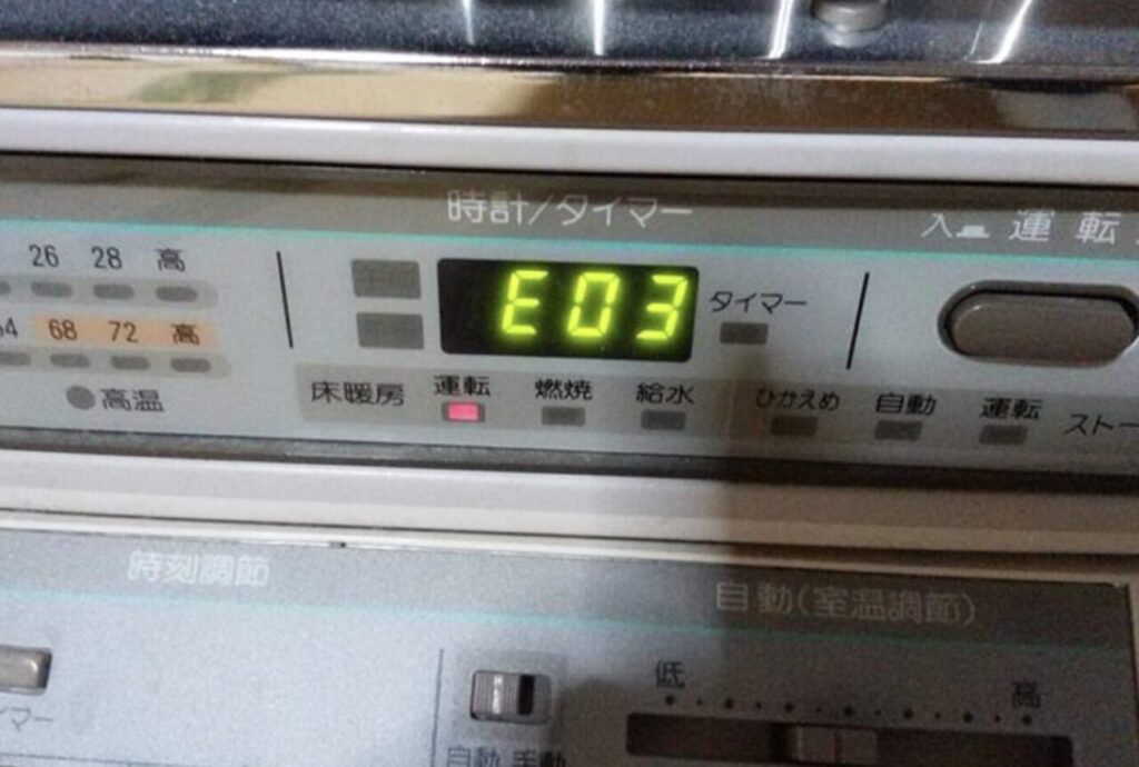 サンポット製の石油ストーブ本体操作パネルに「E03」のエラー表示が点灯している状態。運転ランプが点灯し、床暖房や給水などの表示項目が並ぶ操作部の様子。