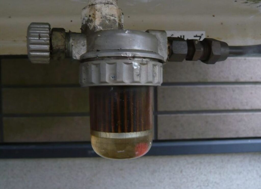 屋外に設置された灯油タンクの下部に取り付けられたストレーナー（灯油フィルター）の様子。透明カップ内に水分や不純物が溜まり、フィルター部の汚れや経年劣化が確認できる状態。