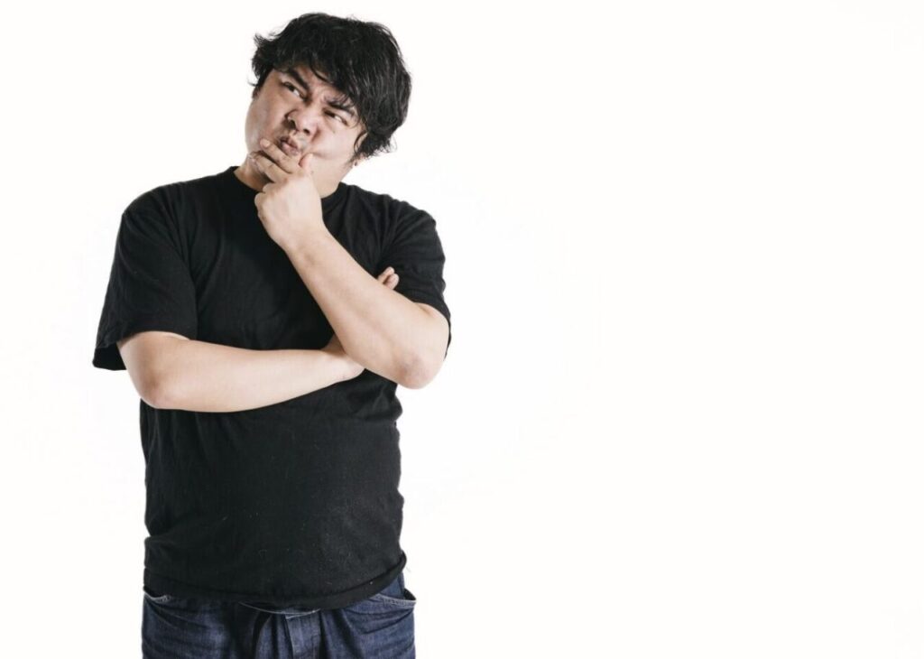 白背景の前で腕を組み、片手を顎に当てて考え込む様子の男性。黒いTシャツを着用し、疑問や迷いを感じている表情が伝わるポーズ。