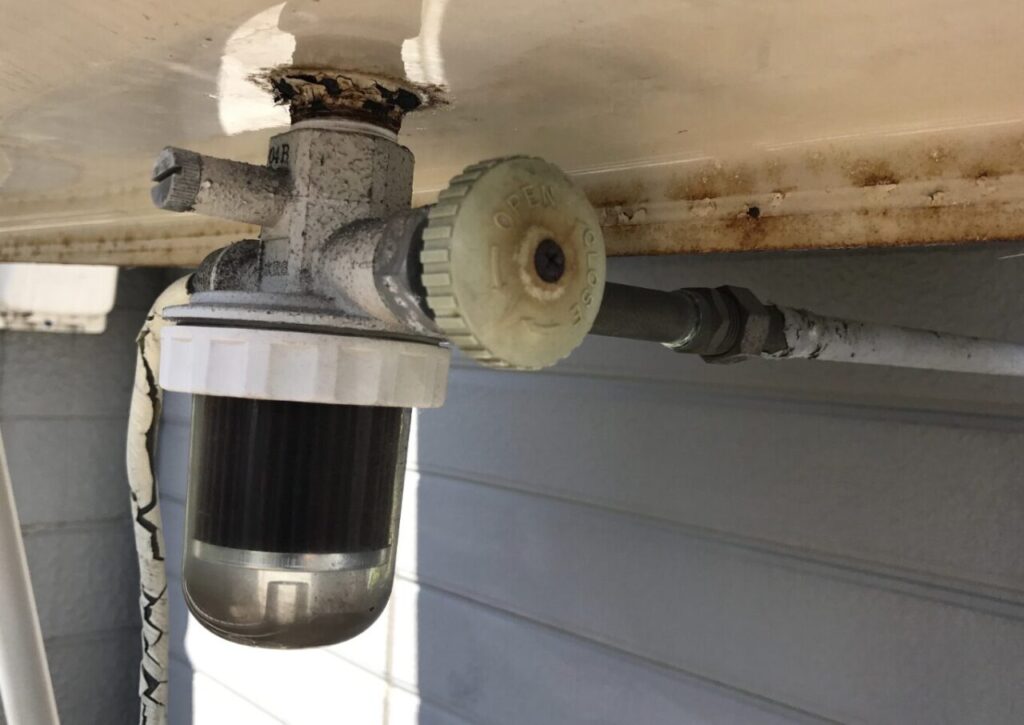 屋外に設置された灯油タンクの下部にあるストレーナー付きバルブの様子。透明カップ内部に灯油の汚れが溜まり、接続部やタンク底面にサビや経年劣化が見られる状態。