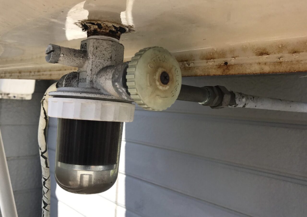 屋外に設置された灯油タンクの下部にあるストレーナー付きバルブの様子。透明カップ内部に灯油の汚れが溜まり、接続部やタンク底面にサビや経年劣化が見られる状態。