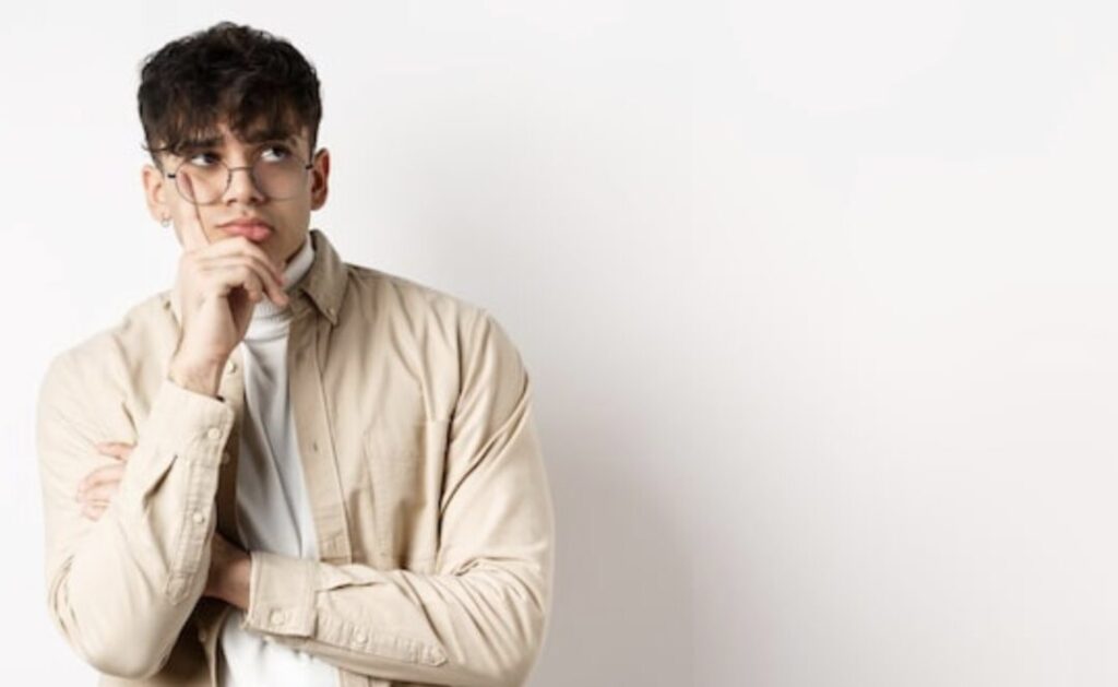 白い背景の前で、眼鏡をかけた男性が腕を組み、顎に手を当てて考え込むような表情をしている様子