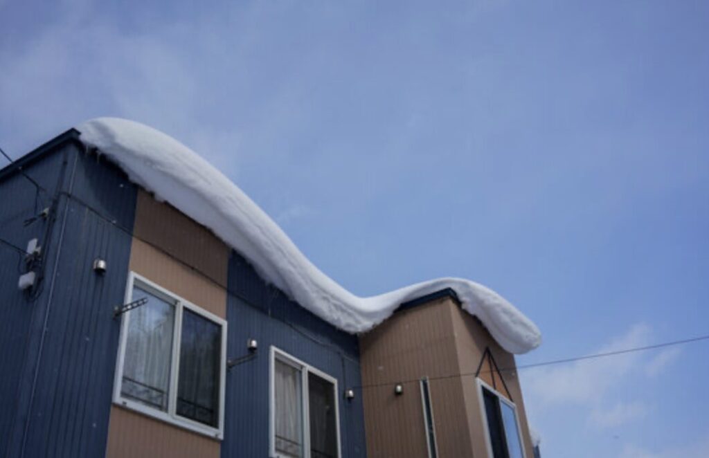 住宅の屋根に大量の雪が積もり、軒先からせり出すように雪庇が形成されている様子を、青空を背景に捉えた冬の住宅外観イメージ。