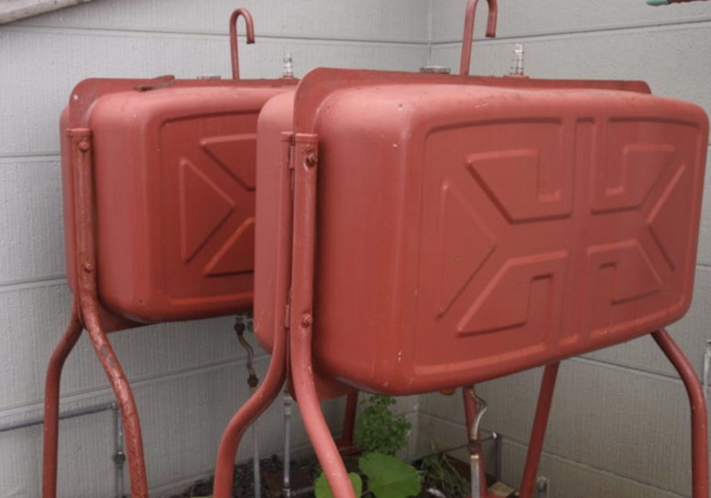 住宅の外壁沿いに設置された赤色の屋外用灯油タンクが2基並んでいる様子。金属製のタンクは専用スタンドで地面から持ち上げられ、給油口や配管が接続された状態で、屋外燃料設備として設置されている状況がわかる構図。