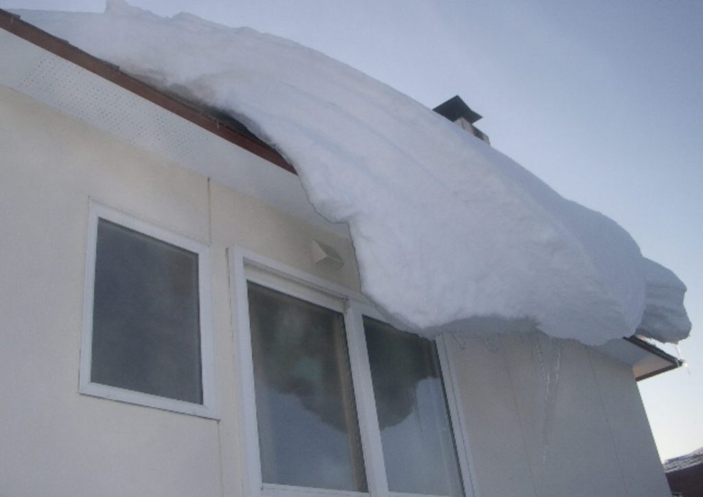 住宅の屋根に積もった雪が軒先から大きく張り出し、外壁上部に設置されたストーブ排気トップ付近まで迫っている外観の様子。